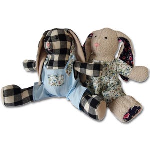 May include: Two plush bunny toys, one wearing blue dungarees with a black and white checked pattern and the other wearing a floral dress with a beige fur body.