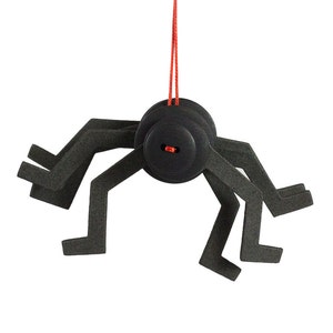 Puede incluir: Una araña de fieltro negro con ocho patas colgando de una cuerda roja. La araña tiene un cuerpo con forma de botón.