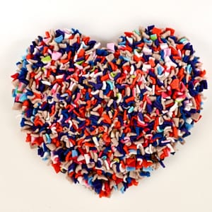 May include: A heart-shaped rug made from colourful fabric scraps. The rug is made of a variety of colours, including red, blue, green, and white.