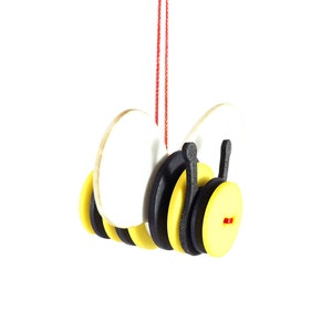 Può includere: Un ornamento di ape in legno con strisce nere e gialle, ali bianche e accenti rossi. L'ape è appesa a uno spago rosso.