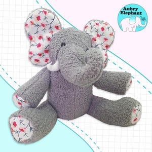 May include: A gray plush elephant toy with pink and white floral fabric ears. The elephant is sitting on a white and blue patterned background.