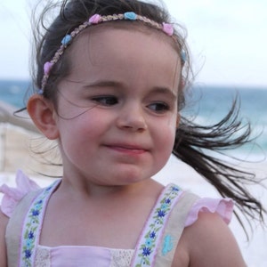 May include: A young girl with long brown hair wearing a pink and blue flower headband and a pink and white dress with floral embroidery.