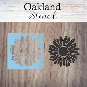 May include: A daisy stencil and its negative space are shown on a light blue square, alongside a black daisy silhouette. The text "Oakland Stencil" is written in a cursive font above the stencils. The background is a wooden surface.