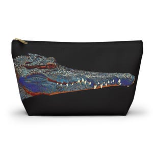 May include: A black zippered pouch featuring a detailed graphic print of a crocodile head in profile. The crocodile is rendered in shades of blue, grey, and orange with visible scales, sharp teeth, and a red eye set against a solid black background.