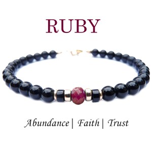Könnte beinhalten: Ein schwarzes Perlenarmband mit einem facettierten Rubin-Edelstein in der Mitte. Das Armband enthält goldfarbene Abstandshalter und einen goldenen Verschluss. Das Wort "RUBY" steht über dem Armband, und die Worte "Fülle | Glaube | Vertrauen" stehen darunter.