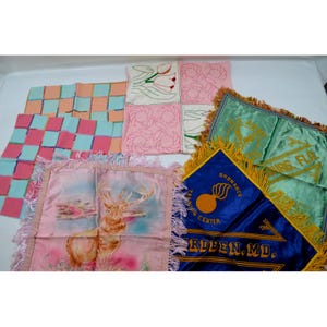 May include: A collection of vintage fabric items including a pink and light blue checkered cloth, a white cloth with embroidered tulips and floral outlines, a green US Army flag with gold fringe, and a blue Ordnance Training Center flag.