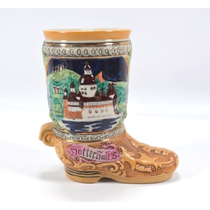 May include: A ceramic boot-shaped stein with a detailed landscape scene featuring a castle, water, and hills. The stein has a tan base, a decorative rim, and the word "Gotterhalt's" on the side.