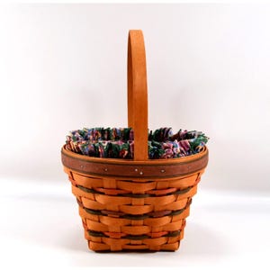May include: A woven basket featuring a tall, arched handle and a tapered body constructed from orange and dark green splints. The rim is accented with a brown band and small metal studs, while the interior is lined with a ruffled floral fabric.
