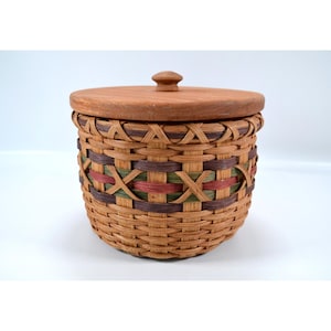 May include: A round woven basket with a wooden lid. The basket is made of light brown woven material with accents of green, red, and brown. The lid is a light brown wood with a small knob on top. The basket is sitting on a white surface.