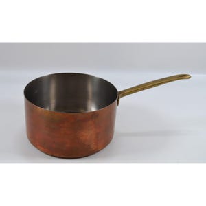 May include: A round copper saucepan with a polished silver-toned interior and a long, straight brass handle attached to the side. The exterior of the pan features a warm, weathered copper finish with visible variations in color and texture.
