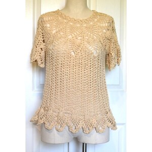 May include: A cream-colored, short-sleeved crochet top. The top features a round neckline and a scalloped hemline. The crochet pattern is open and airy, with a detailed design around the neckline. The top is displayed on a mannequin.