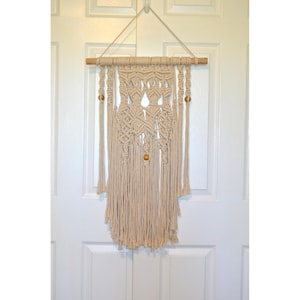 May include: Beige macrame wall hanging with a wooden dowel and wooden beads. The handcrafted textile art features intricate knotting patterns and long fringe, creating a bohemian decorative piece. The wall decor is suspended from a white door.