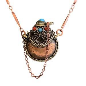 May include: A copper necklace with a decorative pendant featuring a round, ornate, copper container with turquoise and red stones. The pendant is attached to the necklace with a copper chain.