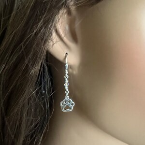 May include: Silver dangle earrings with a paw print charm. The earrings are silver and have a delicate chain.