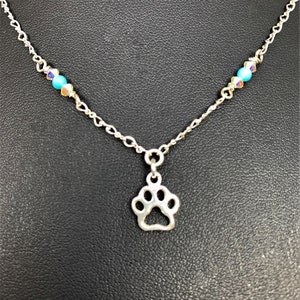 May include: A silver-toned necklace featuring a paw print pendant. The chain is accented with turquoise and iridescent beads. The paw print charm is a cutout design, and the necklace is displayed against a dark background.