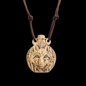 May include: A tan ceramic pendant necklace featuring a detailed wolf face. The pendant is round with carved fur and facial features. It hangs from a brown cord with adjustable knots. The necklace is a unique piece of jewelry.