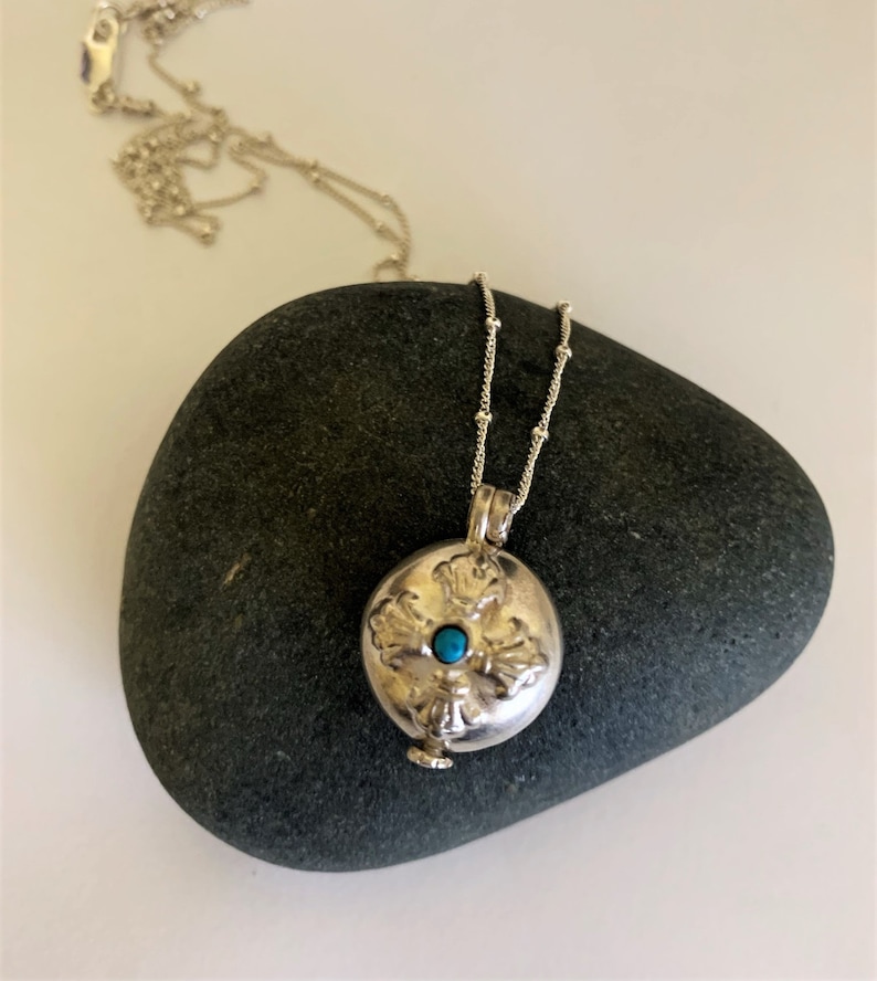 Small round flat silver locket with tiny blue stone in the center. There is a flat round piece at the bottom that is pulled out to open the locket. It sits on a dark gray rock with the chain draped across it. The silver chain drapes across the rock.