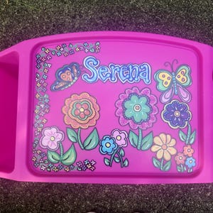 May include: A bright pink plastic lap desk featuring a colorful graphic of flowers, butterflies, and the name Serena written in blue script. The tray includes integrated side compartments for storage and a circular cup holder on the right side.