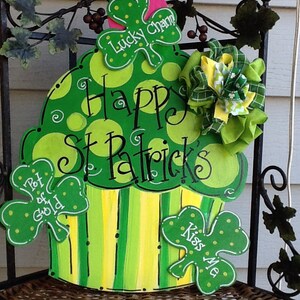 Puede incluir: Un recorte decorativo de madera con forma de cupcake pintado en tonos verdes y amarillos con lunares y rayas. Presenta el texto Happy St. Patrick's, Lucky Charm, Pot of Gold y Kiss Me, decorado con un lazo de tela verde y amarillo.