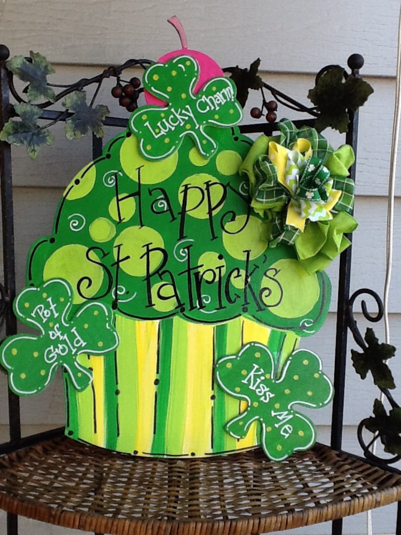 Puede incluir: Un recorte decorativo de madera con forma de cupcake pintado en tonos verdes y amarillos con lunares y rayas. Presenta el texto Happy St. Patrick's, Lucky Charm, Pot of Gold y Kiss Me, decorado con un lazo de tela verde y amarillo.
