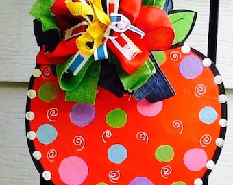 May include: A whimsical, hand-painted wooden pumpkin decoration. The pumpkin is bright orange with colorful polka dots and white swirls. It is topped with a large, multi-colored bow featuring red, green, yellow, and blue ribbons. The edges are outlined in black with white accents.