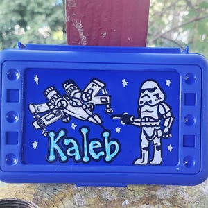 May include: A rectangular blue plastic pencil case featuring a hand-painted design of a white starfighter and a white stormtrooper character. The name Kaleb is written in light blue bubble letters on the front, surrounded by small white stars.