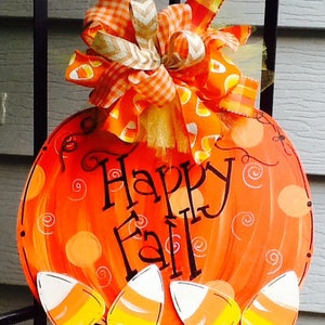 May include: A round orange pumpkin-shaped door sign featuring the black text Happy Fall. The sign is decorated with painted swirls and four candy corn cutouts at the bottom. A large multi-patterned orange and white bow sits at the top.