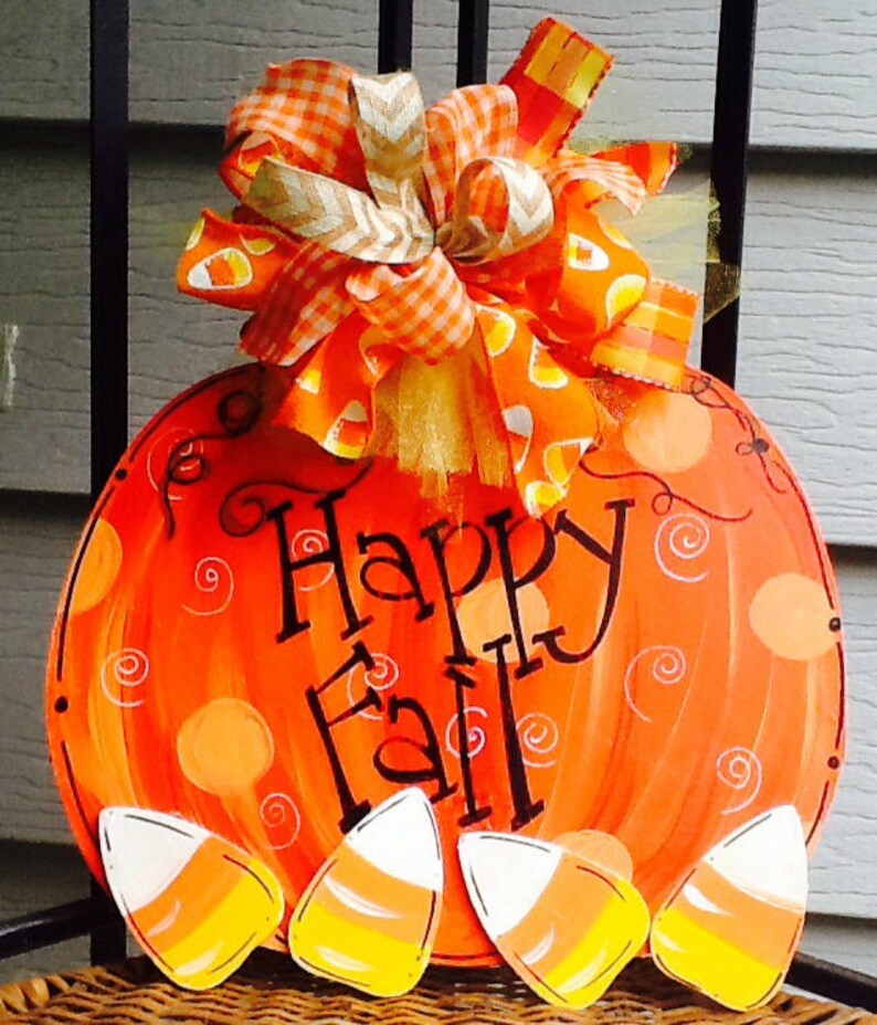 May include: A round orange pumpkin-shaped door sign featuring the black text Happy Fall. The sign is decorated with painted swirls and four candy corn cutouts at the bottom. A large multi-patterned orange and white bow sits at the top.