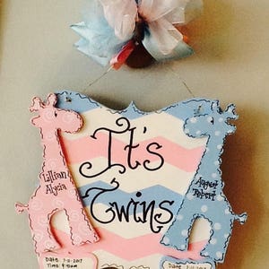 May include: A decorative wooden sign with a pink and blue chevron pattern featuring the text It's Twins. Two giraffe cutouts, one pink and one blue, flank the text. Each giraffe has a small plaque with birth details including date, time, weight, and length.