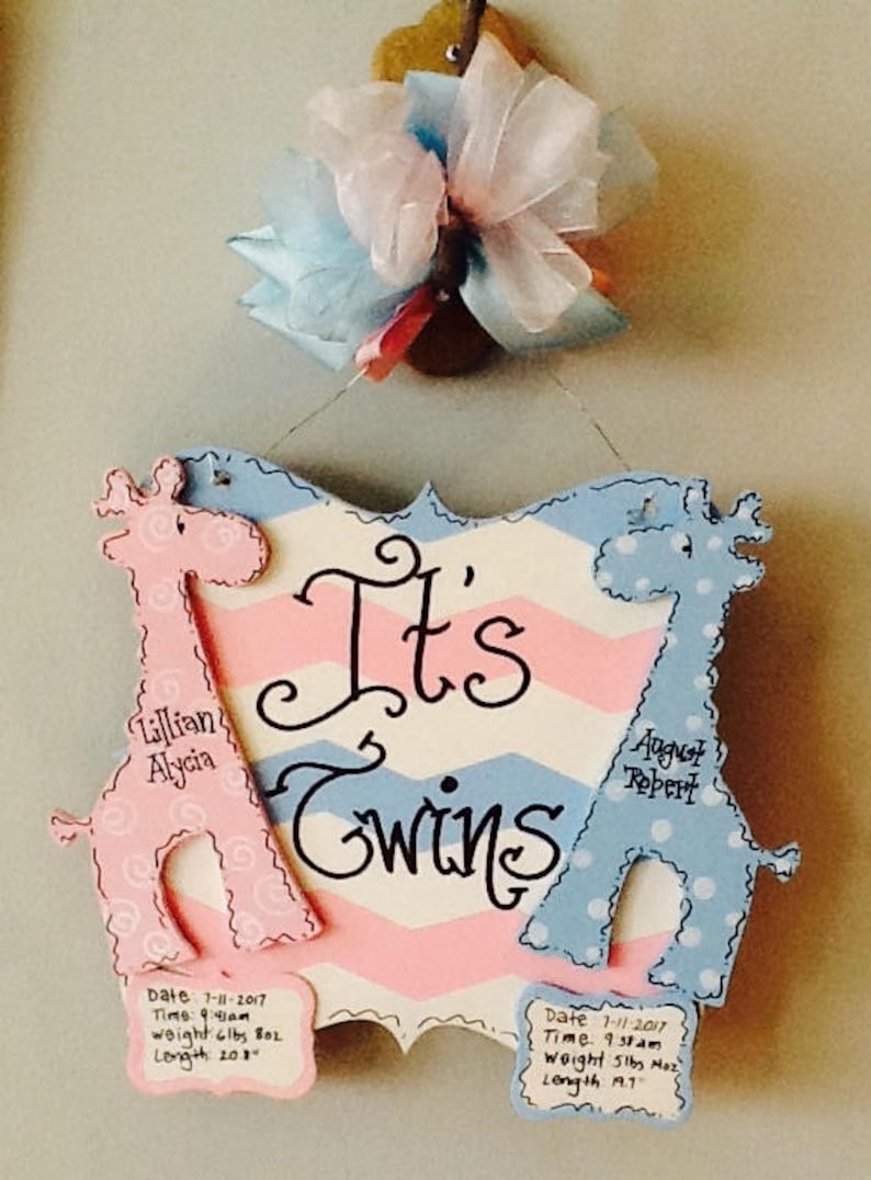May include: A decorative wooden sign with a pink and blue chevron pattern featuring the text It's Twins. Two giraffe cutouts, one pink and one blue, flank the text. Each giraffe has a small plaque with birth details including date, time, weight, and length.