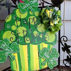 Puede incluir: Un letrero de madera con forma de cupcake verde y amarillo con lunares y rayas. Presenta el texto "Happy St. Patrick's" y tres recortes de tr&eacute;bol etiquetados como "Lucky Charm", "Pot of Gold" y "Kiss Me", acentuados con un lazo de tela.
