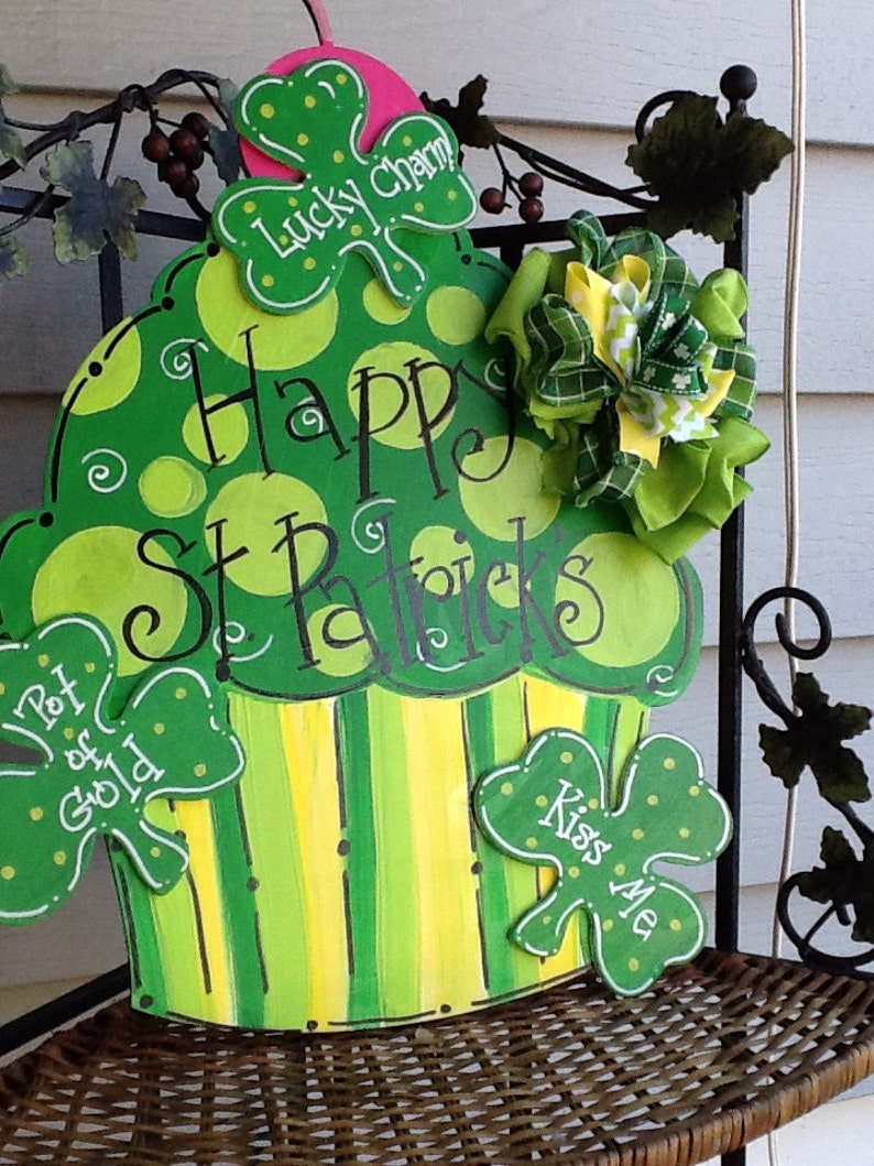 Puede incluir: Un letrero de madera con forma de cupcake verde y amarillo con lunares y rayas. Presenta el texto "Happy St. Patrick's" y tres recortes de tr&eacute;bol etiquetados como "Lucky Charm", "Pot of Gold" y "Kiss Me", acentuados con un lazo de tela.