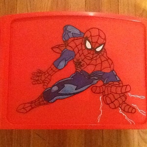 Op de afbeelding: Een felrood plastic schoottafeltje met een centrale grafische illustratie van de superheld Spider-Man in een dynamische gehurkte houding. Het personage is afgebeeld in een rood en blauw pak met een zwart webpatroon en witte ogen.