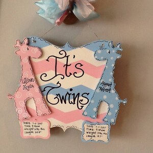 May include: A decorative wooden sign with a pink and blue chevron background featuring the text 'It's Twins'. Two giraffe cutouts, one pink and one blue, flank the text. Each giraffe has a plaque with birth details including date, time, weight, and length.