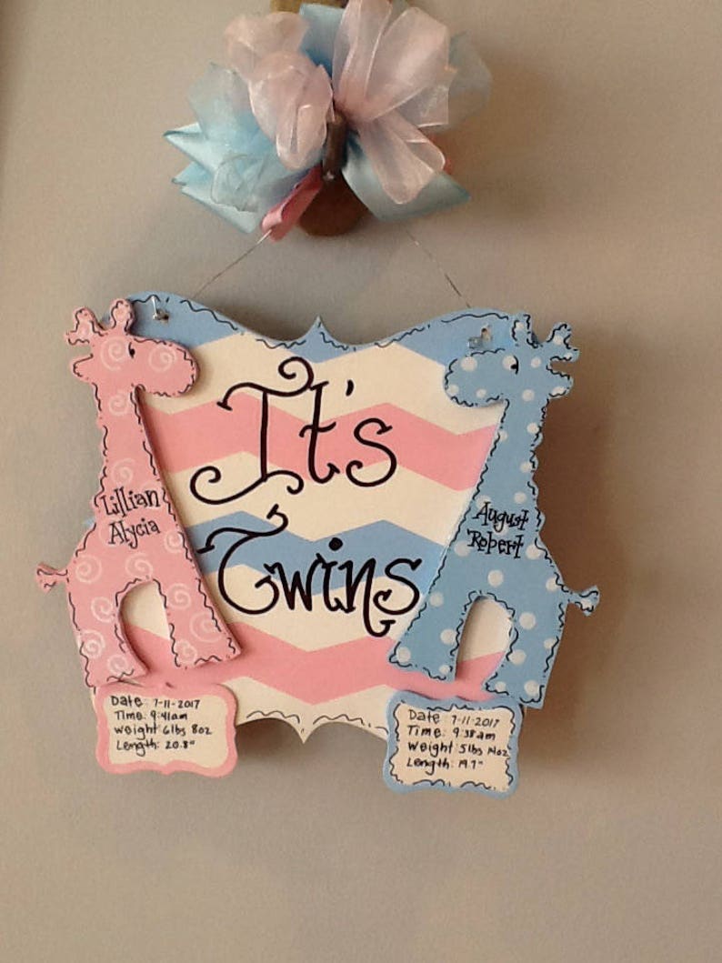 May include: A decorative wooden sign with a pink and blue chevron background featuring the text 'It's Twins'. Two giraffe cutouts, one pink and one blue, flank the text. Each giraffe has a plaque with birth details including date, time, weight, and length.