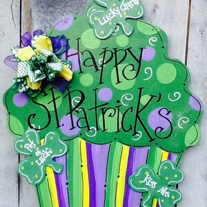 Puede incluir: Un letrero decorativo de madera con forma de cupcake pintado en verde, morado y amarillo. La parte superior presenta lunares verdes y el texto "Happy St. Patrick's". Se incluyen tres recortes de tr&eacute;bol con las etiquetas "Lucky Charm", "Pot of Luck" y "Kiss Me".