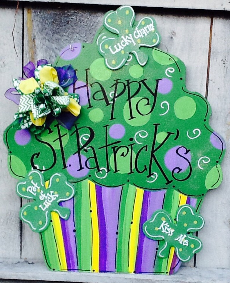 Puede incluir: Un letrero decorativo de madera con forma de cupcake pintado en verde, morado y amarillo. La parte superior presenta lunares verdes y el texto "Happy St. Patrick's". Se incluyen tres recortes de tr&eacute;bol con las etiquetas "Lucky Charm", "Pot of Luck" y "Kiss Me".