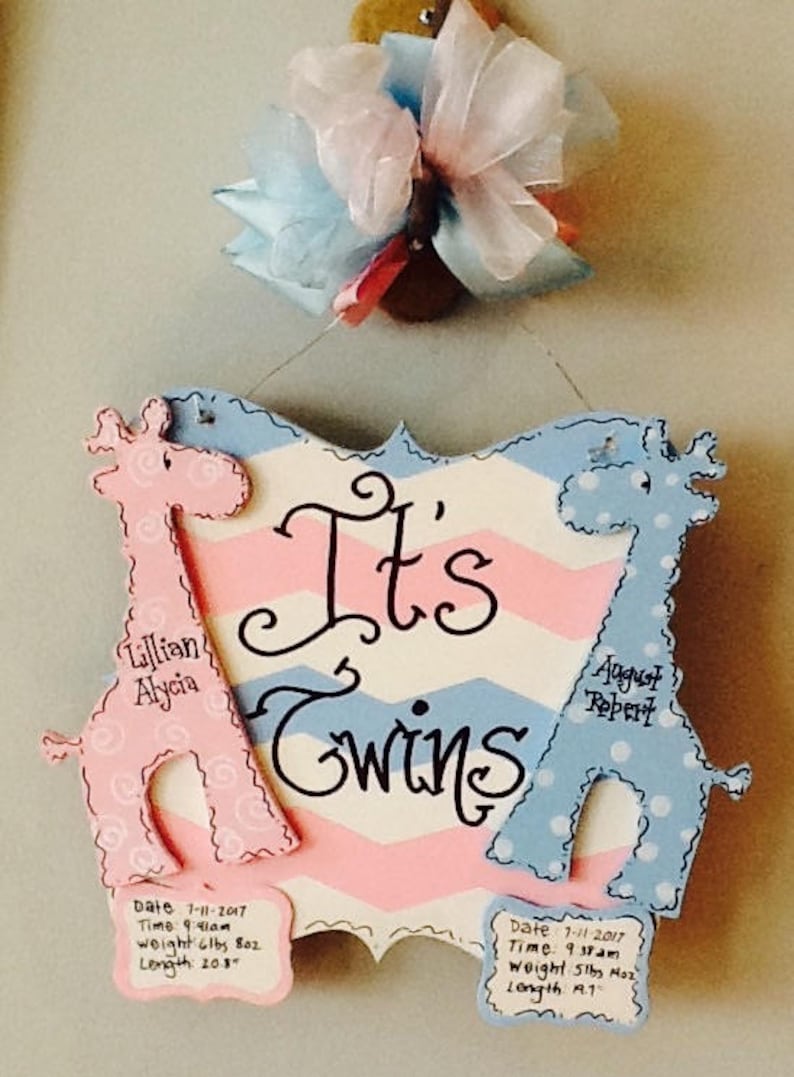 May include: A decorative wooden sign with a chevron pattern in pink, white, and blue, featuring two giraffe cutouts. The text reads It's Twins, with names Lillian Alycia and August Robert, plus birth dates, times, weights, and lengths in inches.
