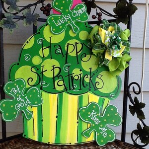 Puede incluir: Un letrero de madera con forma de cupcake verde y amarillo con lunares y rayas. Incluye tres recortes de tr&eacute;bol con el texto Lucky Charm, Pot of Gold y Kiss Me, adem&aacute;s del texto Happy St Patrick's y un lazo de tela verde y amarillo.