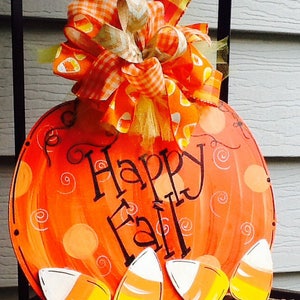 May include: A round orange pumpkin-shaped wooden sign featuring the black hand-painted text Happy Fall. The sign is decorated with painted orange polka dots, swirls, and four candy corn cutouts at the bottom, topped with a large fabric bow.