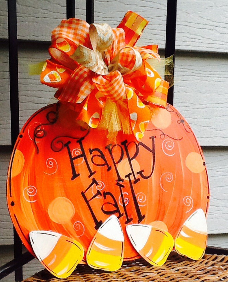 May include: A round orange pumpkin-shaped wooden sign featuring the black hand-painted text Happy Fall. The sign is decorated with painted orange polka dots, swirls, and four candy corn cutouts at the bottom, topped with a large fabric bow.