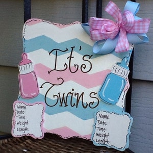 May include: A square wooden sign with a pink, white, and blue chevron pattern featuring the text It's Twins in black script. It includes a pink baby bottle, a blue baby bottle, two blank information cards, and a pink and blue gingham bow.