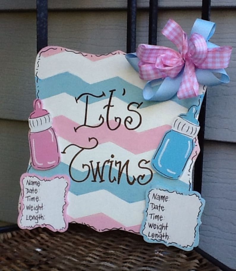 May include: A square wooden sign with a pink, white, and blue chevron pattern featuring the text It's Twins in black script. It includes a pink baby bottle, a blue baby bottle, two blank information cards, and a pink and blue gingham bow.
