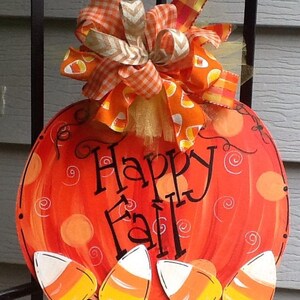 May include: A round orange pumpkin-shaped wooden sign with the text Happy Fall in black script. The sign features painted orange circles and swirls, decorated with four candy corn cutouts at the bottom and a large orange plaid ribbon bow.