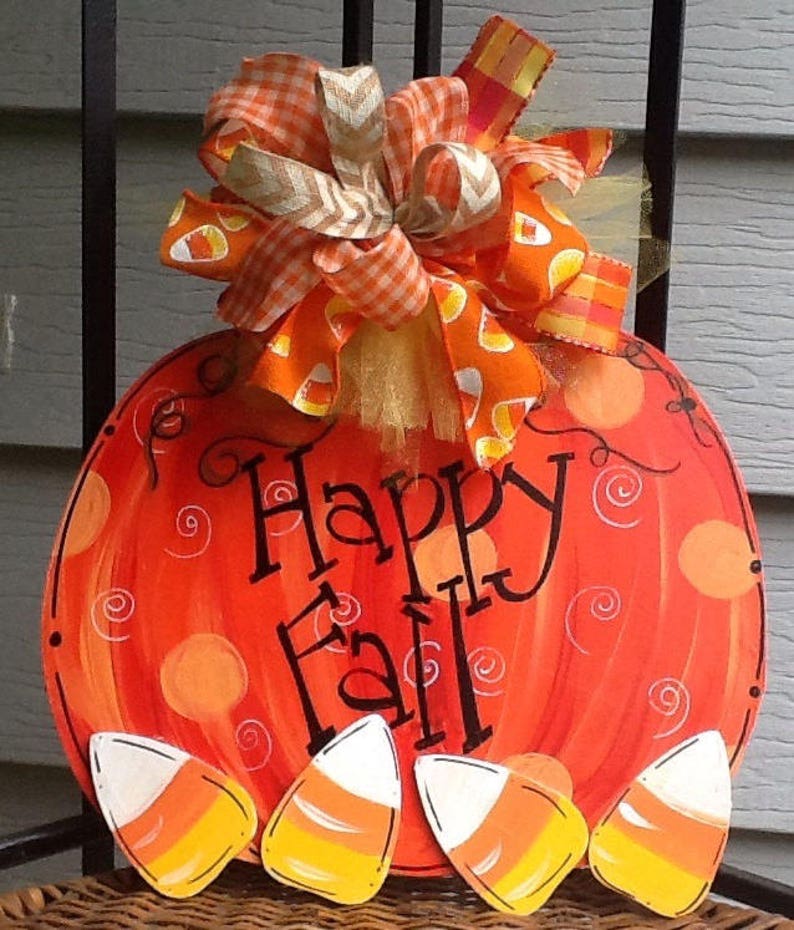 May include: A round orange pumpkin-shaped wooden sign with the text Happy Fall in black script. The sign features painted orange circles and swirls, decorated with four candy corn cutouts at the bottom and a large orange plaid ribbon bow.