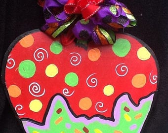 May include: A whimsical, hand-painted cupcake-shaped wooden decoration. The red top features colorful polka dots and white swirls, with a green base adorned with sprinkles. A decorative bow with purple, orange, and red ribbons tops the cupcake.