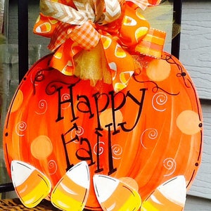 May include: A round orange pumpkin-shaped wooden sign with the text Happy Fall in black script. The sign features painted white swirls and four candy corn cutouts at the bottom. A large decorative bow with orange and white patterns sits on top.