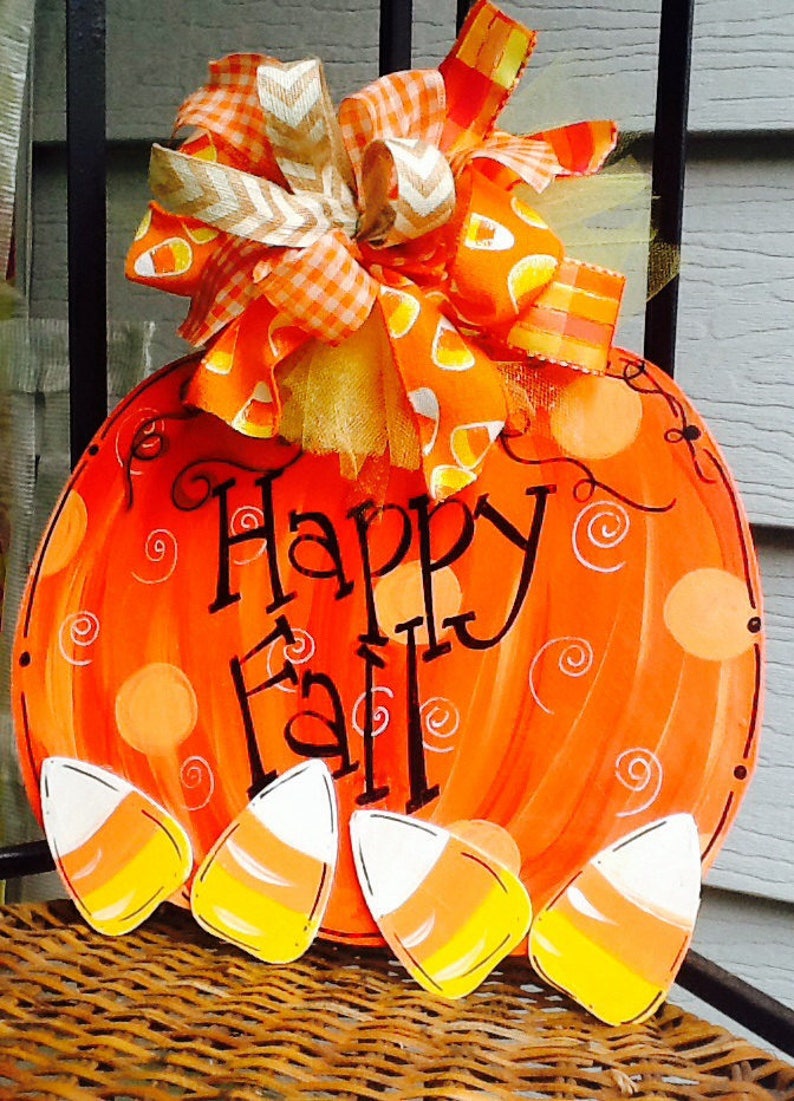 May include: A round orange pumpkin-shaped wooden sign with the text Happy Fall in black script. The sign features painted white swirls and four candy corn cutouts at the bottom. A large decorative bow with orange and white patterns sits on top.