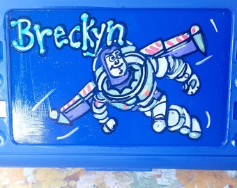 May include: A rectangular blue plastic container featuring a hand-painted illustration of a space ranger character in a white and purple suit with wings. The name Breckyn is written in a stylized blue font above the character on the lid.