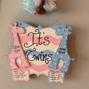 May include: A decorative wooden sign with a pink and blue chevron pattern featuring the text It's Twins. Two giraffe cutouts, one pink with swirls and one blue with dots, flank the text. Each giraffe has a plaque with birth details for Lillian Alycia and August Robert, including dates, times, weights in lbs and oz, and lengths in inches.